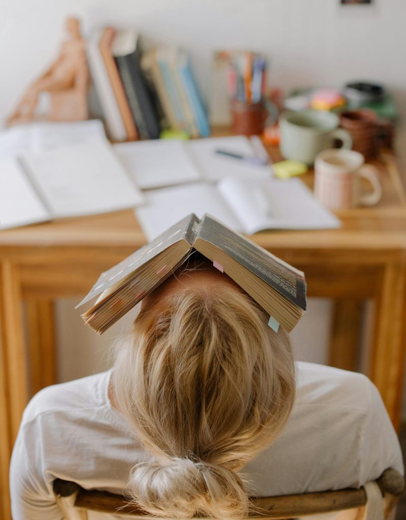 Student feeling stress and exhaustion while studying at a cluttered desk with an open book on their head.